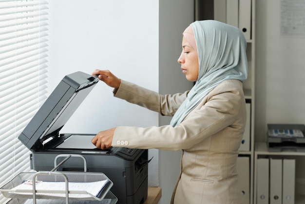 Seitenansicht einer modernen jungen erwachsenen muslimischen Frau mit Hijab, die im Büro arbeitet und Kopien oder Scanni macht