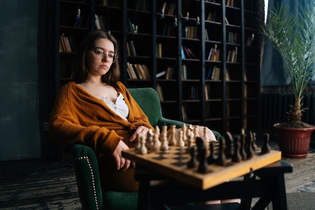 Seitenansicht einer nachdenklichen, ernsthaften jungen Frau mit eleganter Brille, die über Schachzüge nachdenkt und in einem Sessel in einem dunklen Bibliotheksraum sitzt