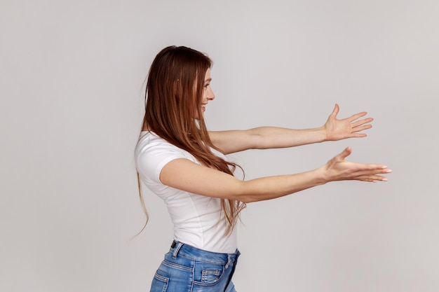 Seitenansicht einer positiven Frau, die mit ausgestreckten Händen kostenlose Umarmungen gibt, einladend einlädt, sich zu umarmen, zu unterstützen und zu pflegen, und ein weißes T-Shirt trägt. Studioaufnahme im Innenbereich isoliert auf grauem Hintergrund.