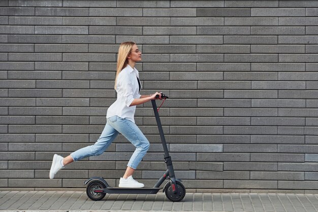 Seitenansicht einer schönen Blondine in Freizeitkleidung, die tagsüber bei sonnigem Wetter mit dem Elektroroller im Freien fährt