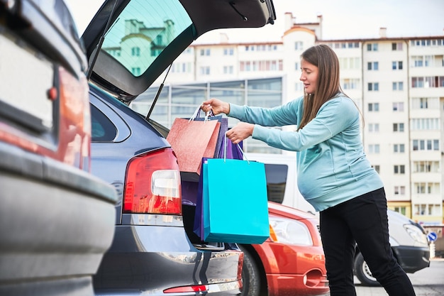 Seitenansicht einer schwangeren Frau mit Bauch, die Einkaufstaschen in den Kofferraum eines Autos legt, die Mutterschaft und Elternschaft im Wartemodus genießt, eine schwangere Frau, die sich während des Schwarzen Freitags auf die Geburt vorbereitet