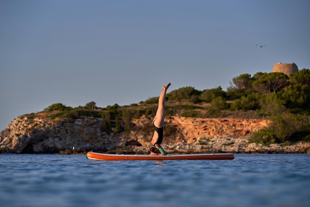 Seitenansicht einer sportlichen Sportlerin im Badeanzug, die Salamba Shirshasana A-Pose auf dem SUP-Board einnimmt, während sie bei Sonnenuntergang Yoga im plätschernden blauen Meer in der Nähe eines felsigen Hügels praktiziert