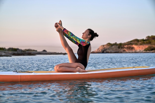 Seitenansicht einer unbeschwerten, sportlichen Frau, die in Heron-Pose meditiert, während sie auf dem Paddleboard inmitten des plätschernden Ozeans Yoga praktiziert