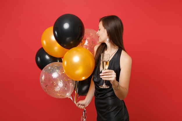 Seitenansicht einer zarten Frau im schwarzen Kleid, die das Halten eines Glases Champagner-Luftballons auf rotem Hintergrund feiert. Internationaler Frauentag Frohes neues Jahr Geburtstag Mockup Urlaub Party Konzept.