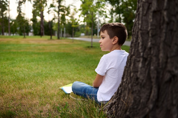 Seitenansicht eines abgelenkten Schuljungen, der sich nach der Schule ausruht, auf dem grünen Gras des Stadtparks sitzt, sich an einen Baum lehnt, seine Hausaufgaben macht und von seiner Umgebung abgelenkt wird. Zurück zur Schule