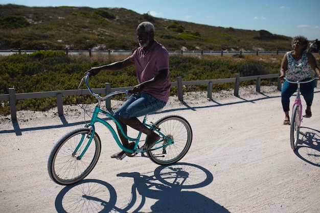 Seitenansicht eines älteren afroamerikanischen Paares, das auf einem Strand Fahrräder fährt, mit blauem Himmel und Hochland im Hintergrund