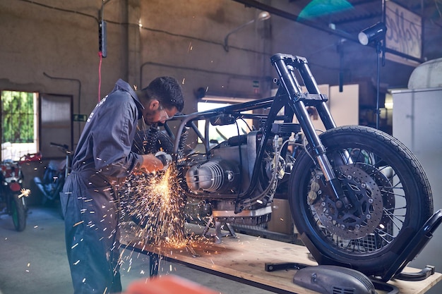 Seitenansicht eines bärtigen Technikers, der Metall mit einem Winkelschleifer schneidet, während er in der Werkstatt ein Motorrad repariert