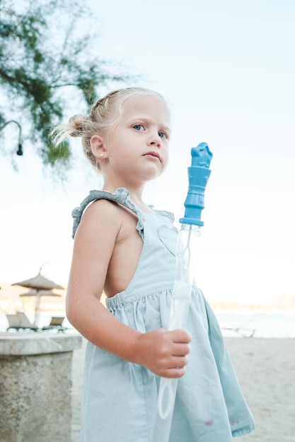 Seitenansicht eines entzückenden kleinen Mädchens im Kleid, das während der Sommerferien eine Plastikflasche mit Seifenblasen in der Hand hält und zum Strand blickt