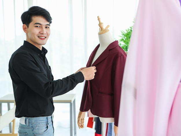 Seitenansicht eines fröhlichen asiatischen männlichen Designers, der während der Arbeit eine stilvolle Jacke auf die Schaufensterpuppe legt.