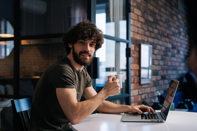 Seitenansicht eines fröhlichen bärtigen jungen Geschäftsmannes, der in der Hand Tasse mit Morgenkaffee hält, der am Tisch mit Laptop-Computer sitzt und in die Kamera blickt