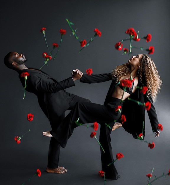 Seitenansicht eines gemischtrassigen barfüßigen Paares von Tänzern, Mann und Frau, die die Augen schließen, in einem stilvollen schwarzen Anzug tragen und Händchen halten und sich zurücklehnen, während rote Blumen von oben fallen