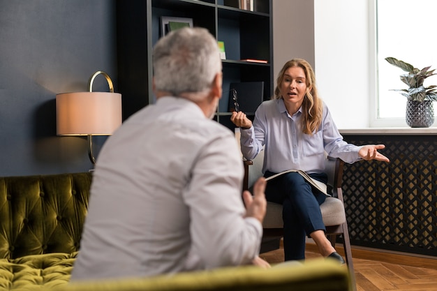 Seitenansicht eines grauhaarigen Mannes, der auf dem olivgrünen Sofa sitzt, während er einer blonden Frau zuhört, die mit ihm spricht