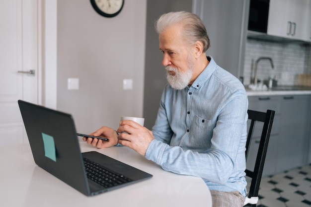 Seitenansicht eines grauhaarigen, reifen erwachsenen Geschäftsmannes, der am Laptop arbeitet und sein Mobiltelefon mit einer Tasse Kaffee am Tisch hält