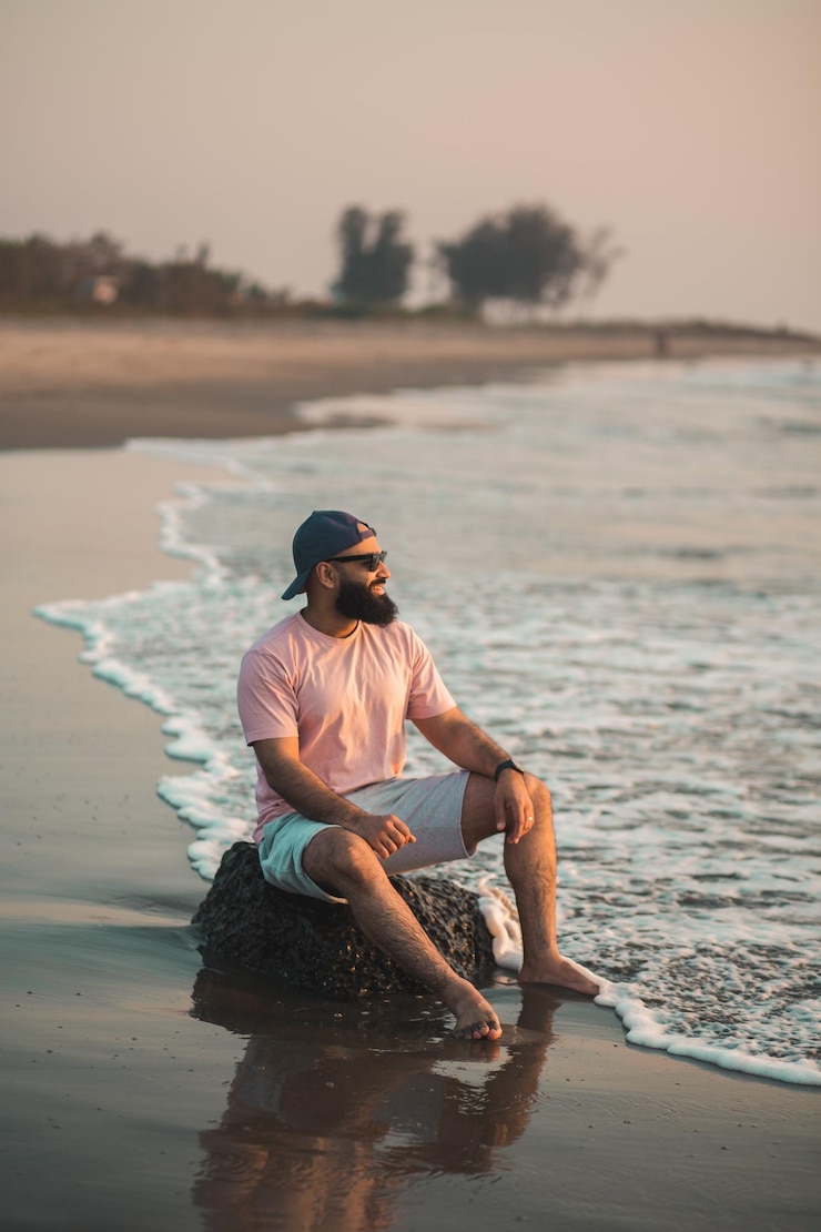 Foto seitenansicht eines gut aussehenden bärtigen mannes, der bei sonnenuntergang mit den füßen im wasser am strand sitzt