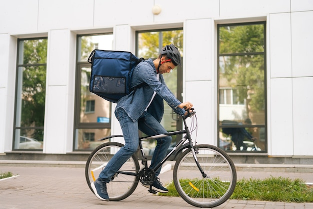 Seitenansicht eines gutaussehenden jungen Liefermannes in Schutzhelm, der eine Fahrradtour in der Stadtstraße auf dem Hintergrund des Bürogebäudes beginnt. Kuriermännchen mit Thermorucksack, der Essen an den Kunden liefert