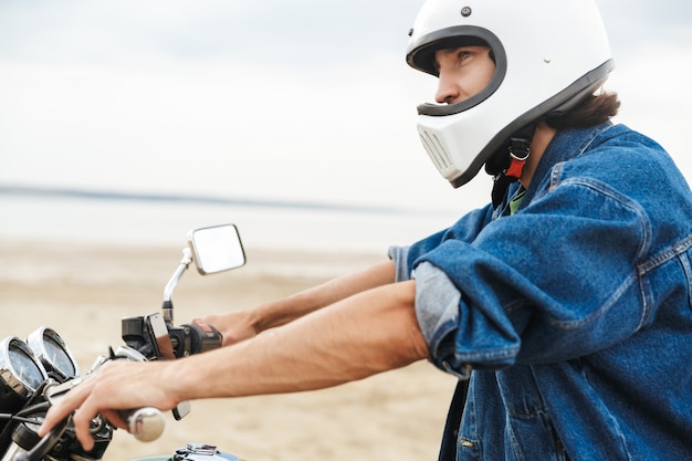 Seitenansicht eines gutaussehenden jungen Mannes in lässigem Outfit, der auf einem Motorrad am Strand sitzt und einen Helm trägt