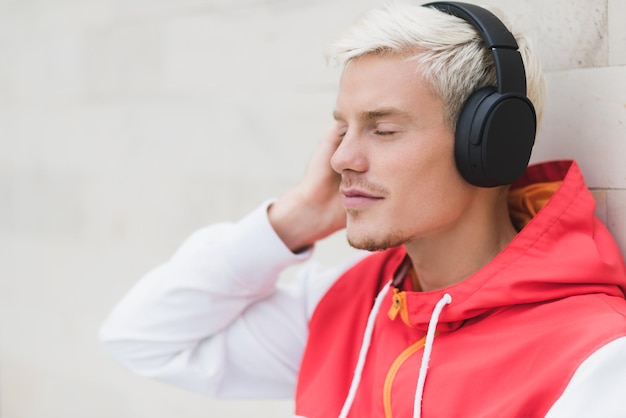 Seitenansicht eines gutaussehenden Mannes mit blonden Haaren in roter Sportbekleidung, der sich nach körperlichen Aktivitäten an der grauen Wand draußen entspannt und sitzt Stilvoller Mann, der Musik über Bluetooth-Kopfhörer hört