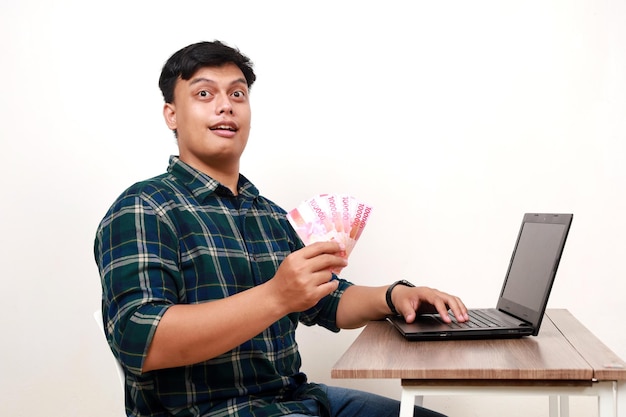 Seitenansicht eines jungen asiatischen College-Studenten, der sitzt, während er indonesische Banknoten hält und einen Laptop auf dem Schreibtisch benutzt