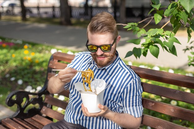 Seitenansicht eines jungen gutaussehenden Hipster-Mannes, der chinesische Nudeln von einer Brotdose isst, während er in einem sitzt