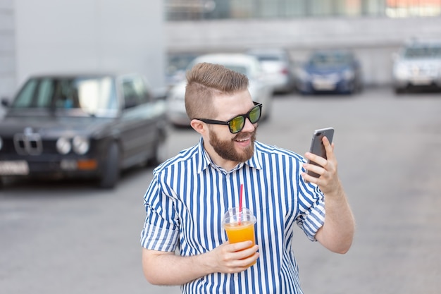 Seitenansicht eines jungen stilvollen Hipster-Mannes mit Brille und einem Schnurrbart und Saft in seinen Händen, die in den sozialen Netzwerken unter Verwendung eines Smartphones und des drahtlosen Internets an einem warmen Sommertag plaudern.