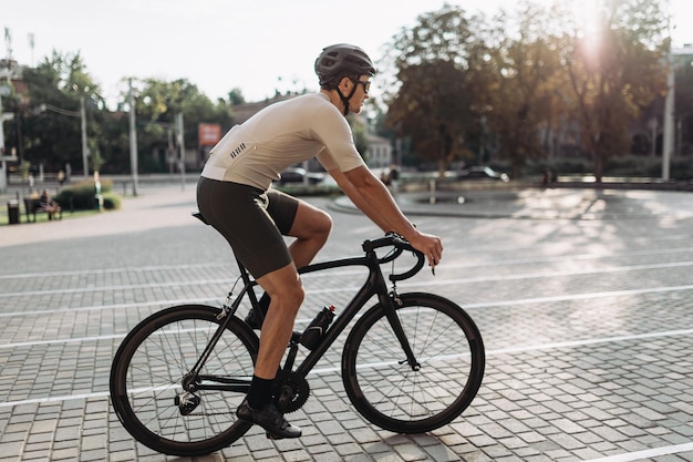 Seitenansicht eines kaukasischen Mannes mit muskulösen Beinen, der ein schwarzes Fahrrad auf der Straße der Stadt fährt Erfahrener Radfahrer mit Sportkleidung, Schutzhelm und verspiegelter Brille