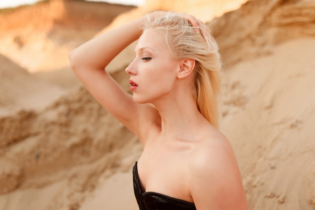 Seitenansicht eines kaukasischen weiblichen Modells mit blondem Haar und Make-up, im schwarzen sexy Bodysuit, ihr Haar berührend, auf dem Sand in der Wüste sitzend.