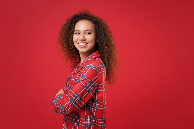 Seitenansicht eines lächelnden afrikanisch-amerikanischen Mädchens in Pyjama-Homewear, das zu Hause isoliert auf rotem Hintergrund ruht. Entspannen Sie sich bei guter Laune Lifestyle-Konzept. Mock-up-Kopienbereich. Händchen haltend gekreuzt, zur Seite schauend.