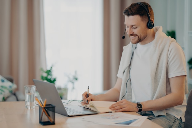 Seitenansicht eines lächelnden Mannes im Headset, der Online-Videounterricht hat und während des E-Learning zu Hause einige Notizen im Notizbuch schreibt. Menschen und Fernbildung