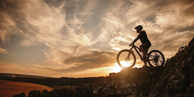 Seitenansicht eines männlichen Radfahrers im Helm, der auf dem Mountainbike gegen den bewölkten Sonnenuntergangshimmel in der Landschaft sitzt