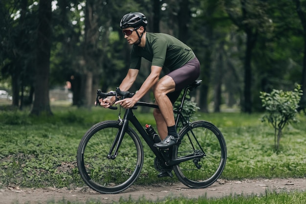 Seitenansicht eines männlichen Sportlers in Sportkleidung, Schutzhelm und verspiegelter Brille, der im Sommerpark Fahrrad fährt. Konzept der Ausdauer, Selbstdisziplin und Motivation.