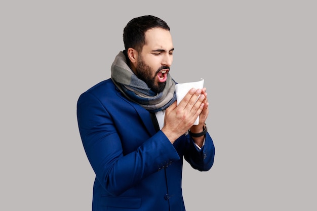 Seitenansicht eines Mannes, der in einen warmen Schal gehüllt ist und in einer Serviette niest, die eine laufende Nase reinigt, die an Grippesymptomen leidet