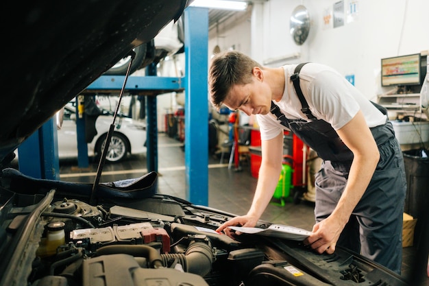 Seitenansicht eines professionellen gutaussehenden professionellen männlichen Automechanikers in blauer Uniform, der vor offener Motorhaube steht und ein Klemmbrett hält, das den Motor des Autos untersucht, das zur Reparatur oder Wartung ankommt.