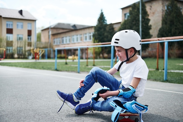 Seitenansicht eines sportlichen Jungen im Schutzhelm, der auf einem Holzskateboard sitzt und schützende Knieschützer anzieht