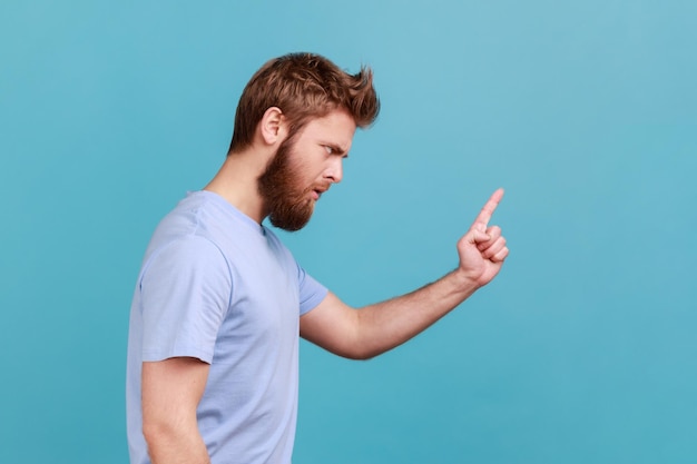 Seitenansicht eines strengen, ernsthaften jungen bärtigen Mannes, der vor Fehlern warnt, der mit mahnendem Finger auf den Seitenraum schaut und alarmierend Ratschläge gibt Studioaufnahme im Innenbereich isoliert auf blauem Hintergrund