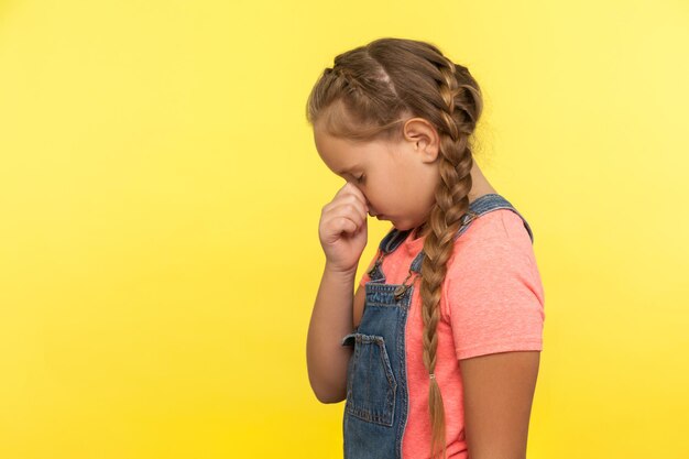 Seitenansicht eines unglücklichen kleinen Mädchens mit Zopf in Denim-Overalls, das sich mit dem Gesicht nach unten versteckt und weint, verärgert über Verlust oder Niederlage, Kind, das Trauer und Bedauern empfindet. Studioaufnahme im Innenbereich isoliert auf gelbem Hintergrund