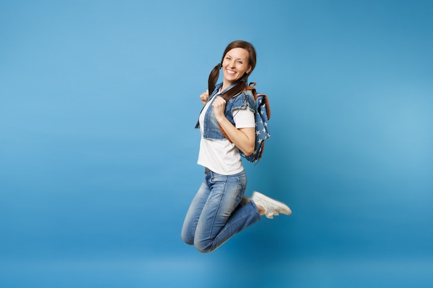 Seitenansicht Ganzaufnahme der jungen fröhlichen glücklichen Studentin in Denimkleidung mit Rucksackspringen einzeln auf blauem Hintergrund. Ausbildung an der Fachhochschule. Kopieren Sie Platz für Werbung.