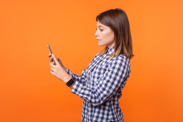 Seitenansicht Porträt einer ernsthaften, schönen jungen Frau mit braunen Haaren, die ein kariertes Hemd trägt, das mit einem Handy steht, das einen Videoanruf tätigt oder eine Studioaufnahme im Innenbereich bloggt, isoliert auf orangefarbenem Hintergrund