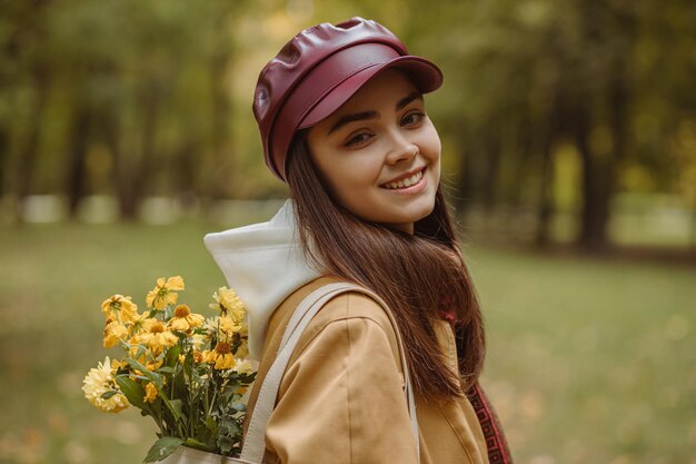 Seitenansicht Porträt einer fröhlichen Frau mit Blumenstrauß in der Tasche, die im Herbst im Park in die Kamera blickt