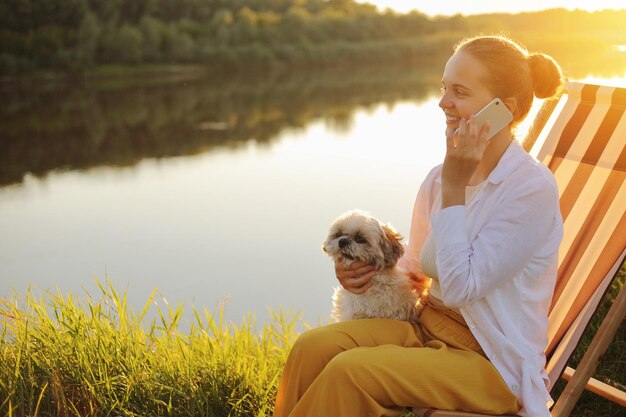 Seitenansicht Porträt einer glücklichen positiven Frau mit weißem Hemd, die mit ihrem pekinesischen Hund in der Nähe des Flusses auf einem Liegestuhl sitzt und mit dem Handy telefoniert und ein angenehmes Gespräch führt