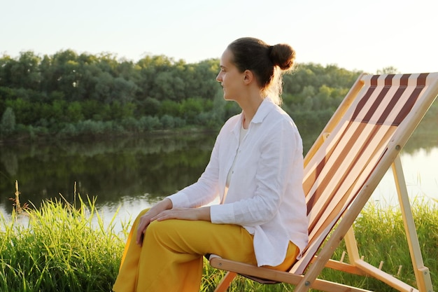 Seitenansicht Porträt einer jungen erwachsenen Frau, die im Klappstuhl am malerischen Ufer des Flusses sitzt und die Ruhe der Sommernatur genießt und die Freiheit der Tiefe beobachtet