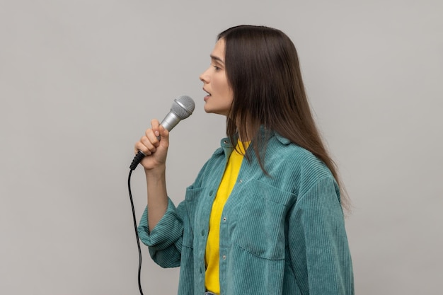 Seitenansicht Porträt einer jungen erwachsenen Journalistin, die ein Mikrofon hält und Probleme bespricht, die das Tragen einer Jacke im legeren Stil melden Innenstudioaufnahme isoliert auf grauem Hintergrund