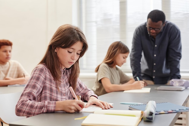 Seitenansicht Porträt einer jungen Teenagerin, die in ein Notizbuch schreibt, während sie am Schreibtisch im Kopierraum der Schulklasse sitzt