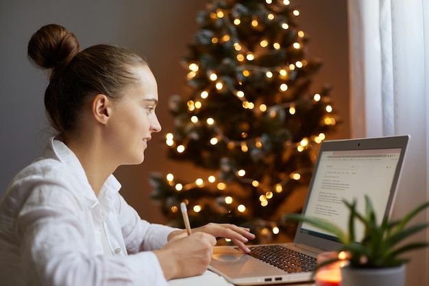 Seitenansicht Porträt einer kaukasischen Frau, die zu Hause aus der Ferne arbeitet, mit einem Laptop, der am Tisch sitzt und in ein Notizbuch aus Papier schreibt, das auf dem Hintergrund eines Weihnachtsbaums posiert