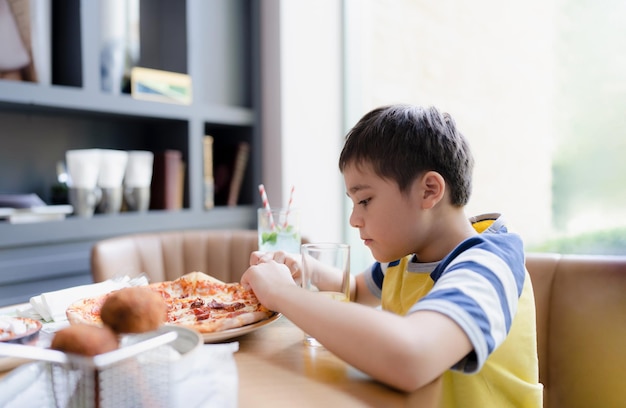 Seitenansicht Porträt eines Kindes, das im Café hausgemachte Pizza isst. Glücklicher Kinderjunge, der im Restaurant ein großes Stück frisch zubereitete Pizza abbeißt. Familienglückliches Zeitkonzept