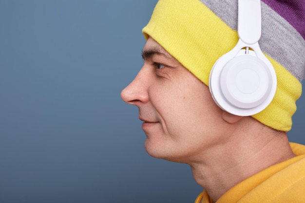 Seitenansicht Porträt eines ruhigen, lächelnden, attraktiven Mannes mit Mütze und lässigem gelbem Hoodie, der an der blauen Wand posiert und auf den Kopierraum für Werbung wegblickt, die Musik über Kopfhörer hört