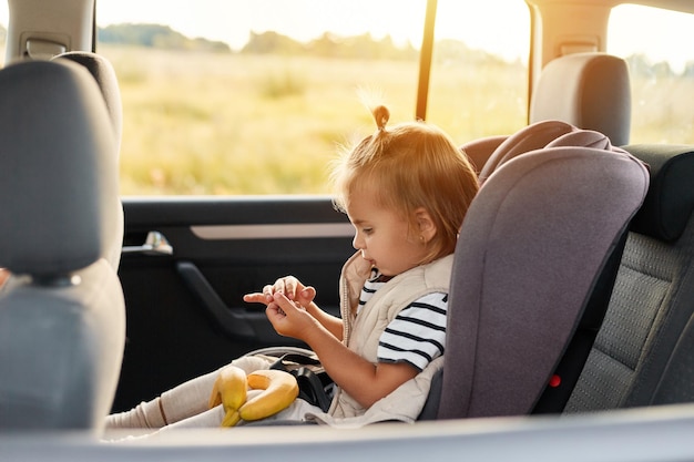 Seitenansicht Porträt eines süßen, charmanten Mädchens, das in einem Auto auf einem Sicherheitsstuhl sitzt und alleine spielt und Bananen hält, die ein gestreiftes T-Shirt auf Reisen tragen
