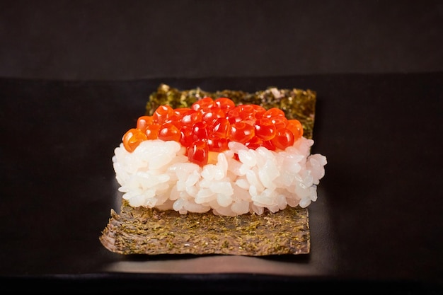 Seitenansicht von Ikura Sushi Lachsrogen-Sushi auf getrockneten Algen auf einem schwarzen Teller platziert, isoliert auf schwarzem Hintergrund beliebtes japanisches Essen