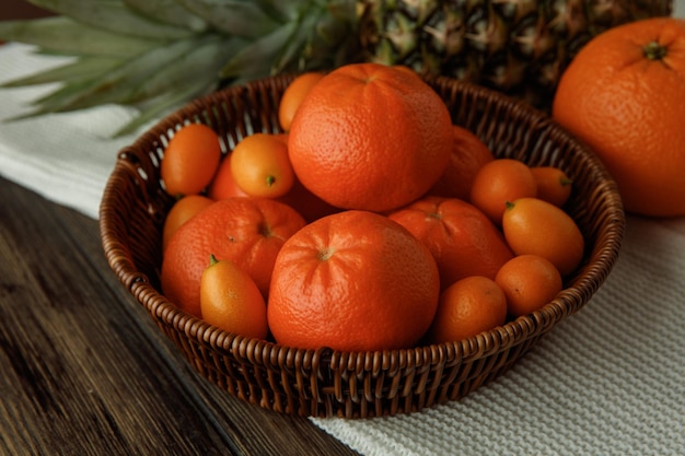 Seitenansicht von Zitrusfrüchten als Mandarinen und Kumquats in Korborangenananas auf Stoff auf Holzhintergrund