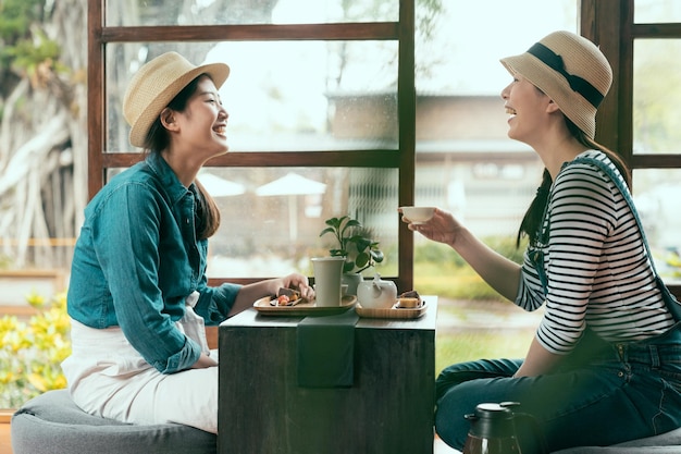 seitenansicht von zwei asiatischen freundinnen, die drinnen im lokalen restaurant von kyoto lachen und plaudern. glückliche schwestern, die während der teezeremonie einen snack essen und tee trinken. Damen genießen die Nachmittagsfreizeit im Garten