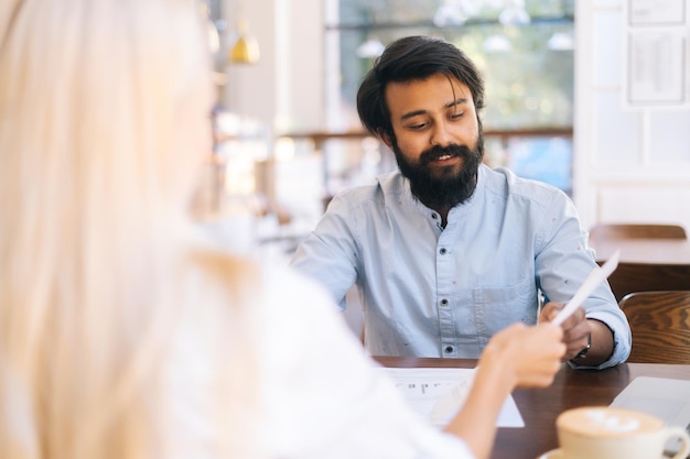 Seitenansicht von zwei multiethnischen Kollegen diskutieren Finanzdaten in Berichten mit Dokumenten und Diagrammen, die am Schreibtisch im Café am Fenster sitzen